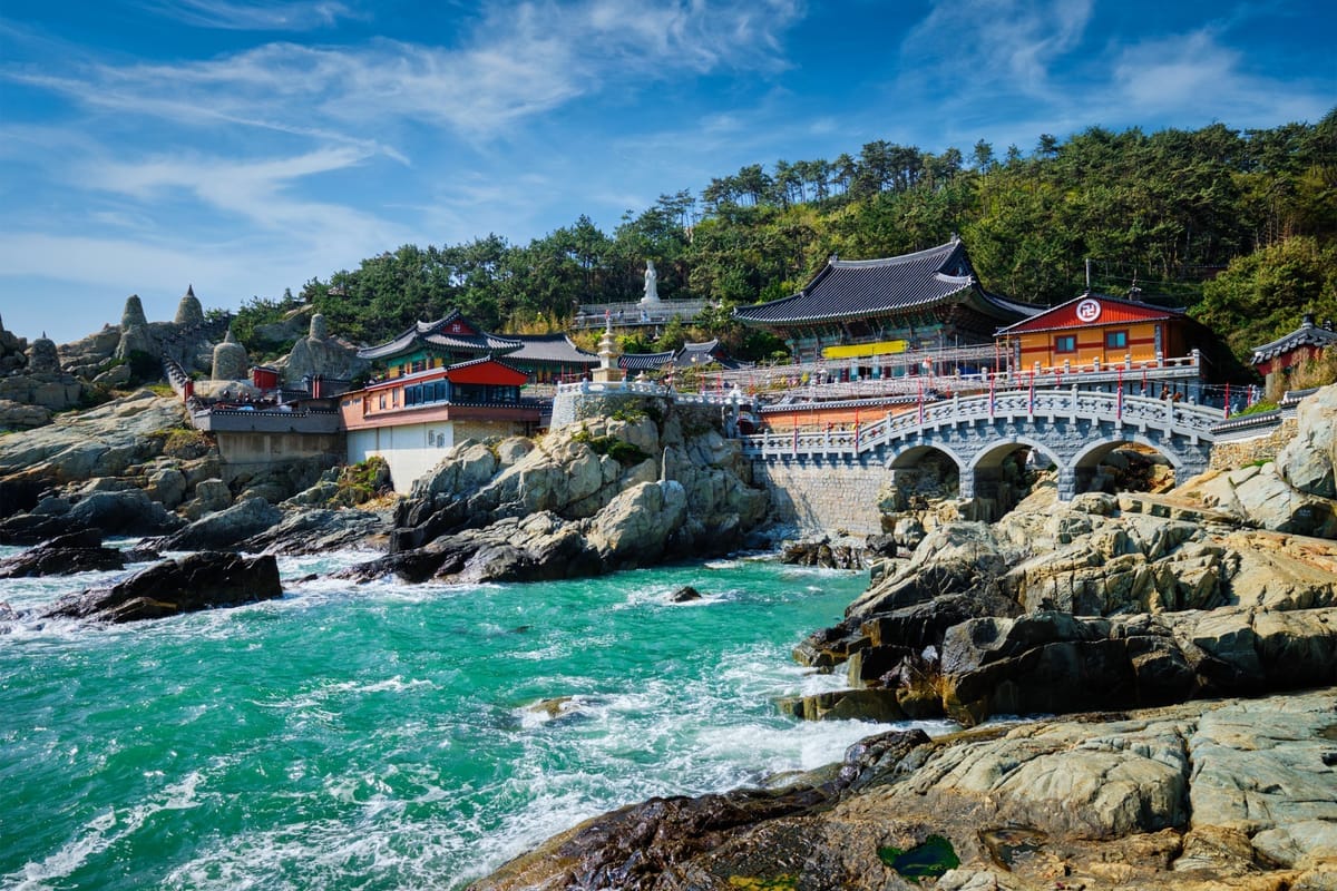 Der Tempel Haedong Yonggungsa mit roten und goldenen Hallen über der felsigen Küste bei Busan, davor eine weiße Steinbrücke über die Brandung