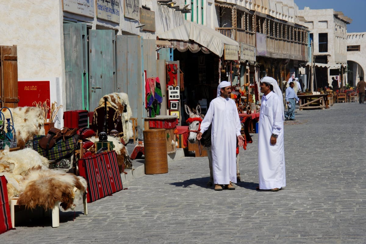 Gepflasterte Gasse im Souq Waqif mit Lehmbauten, ausgelegten Teppichen und Fellen vor den Läden, im Hintergrund zwei Männer in weißen Thawbs im Gespräch
