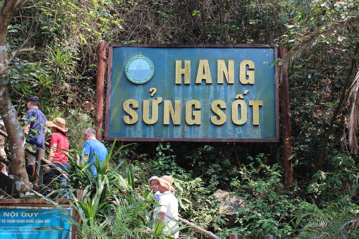 Blaues Hinweisschild „Hang Sung Sot“ zwischen dichtem Grün, davor Besucher auf der Treppe zum Höhleneingang