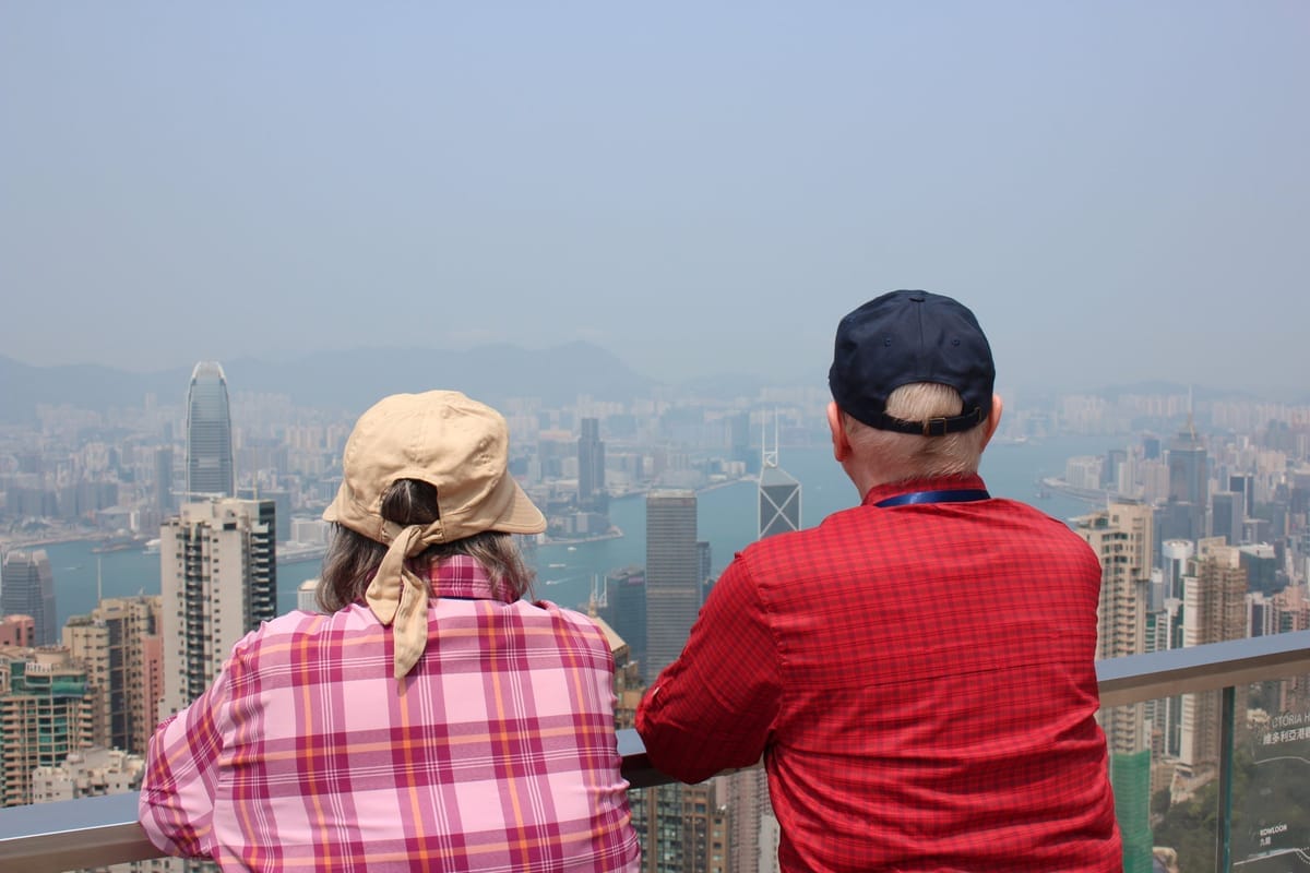 Zwei Besucher stehen an der Brüstung einer Aussichtsplattform auf dem Victoria Peak und blicken über die Hochhäuser von Hongkong Island zum Victoria Harbour