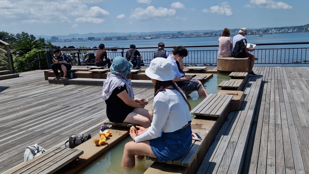 Besucher sitzen mit den Füßen im warmen Wasser des Fußbads auf Sakurajima, dahinter die Bucht und die Stadt Kagoshima