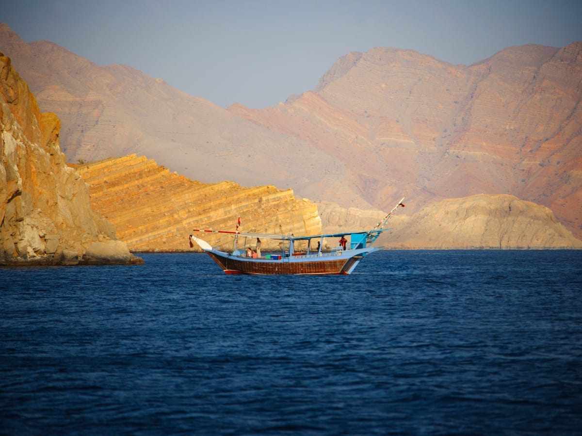 Traditionelle hölzerne Dhau auf ruhigem Wasser vor einer senkrecht aufragenden Felswand der Musandam-Halbinsel