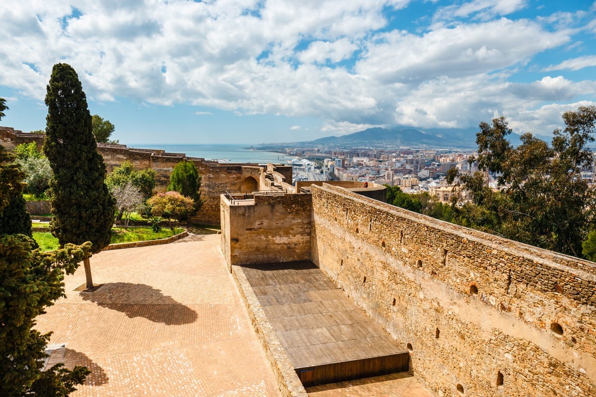 Verwitterte Festungsmauern der Alcazaba von Málaga, die sich einen Hang hinaufziehen, dahinter Stadtbebauung