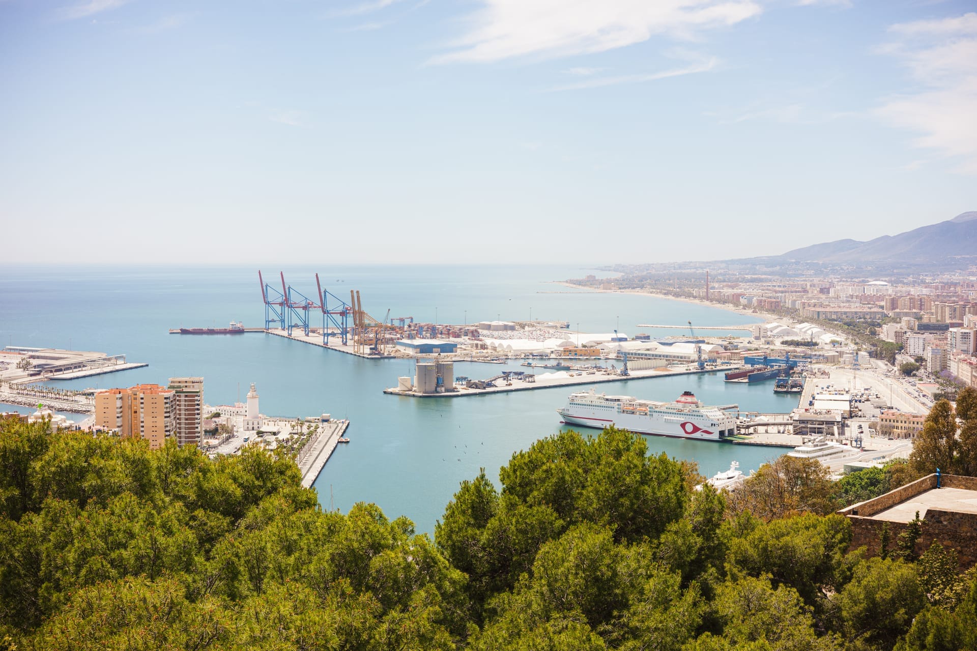 Blick von oben über Málaga mit Hafenbecken, Kränen und Schiffen, dahinter die Stadt und die Küste
