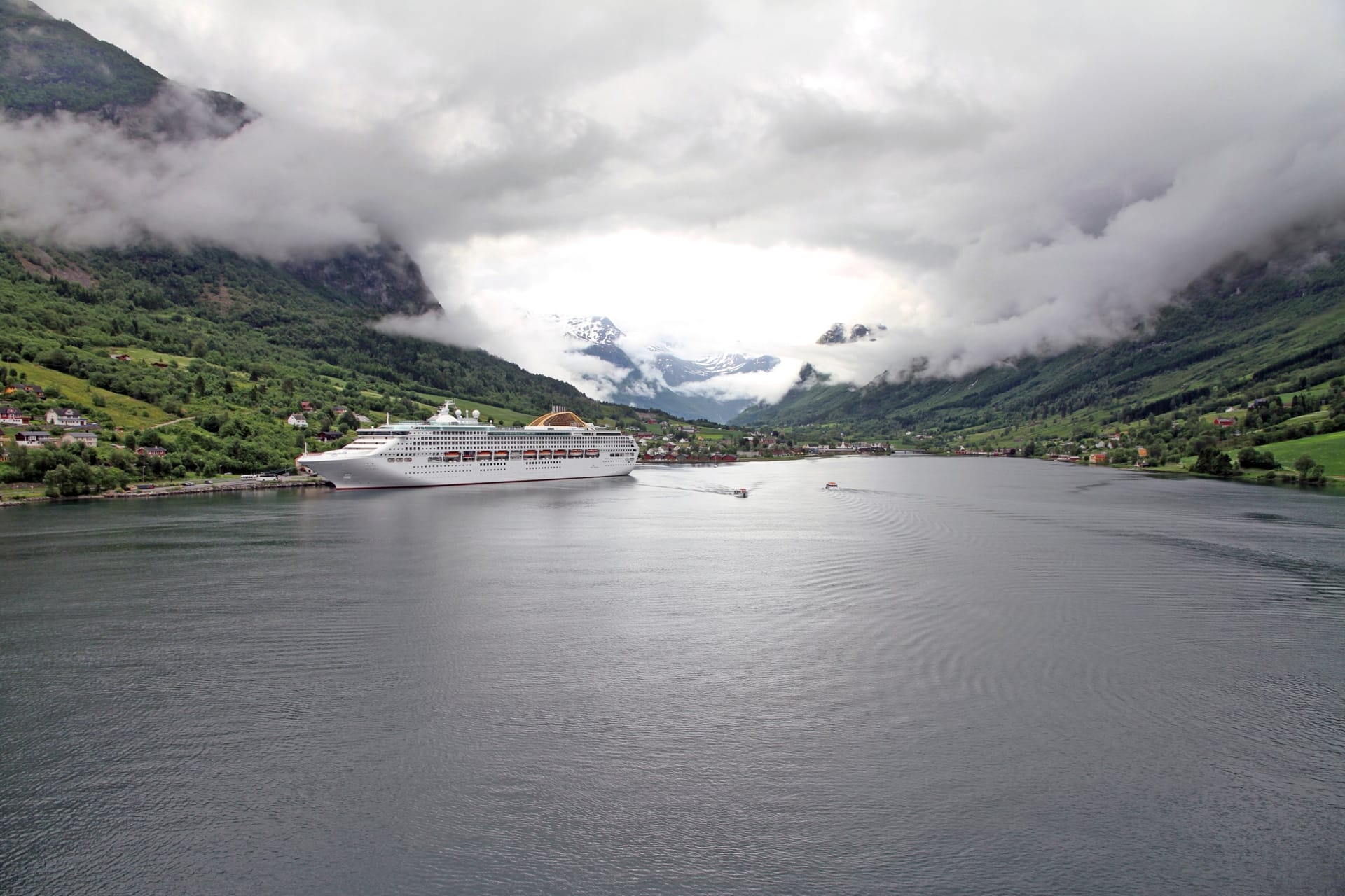 Kreuzfahrtschiff im Nordfjord vor dem Ort Olden, die grünen Berghänge in tief hängenden Wolken, dahinter schneebedeckte Gipfel