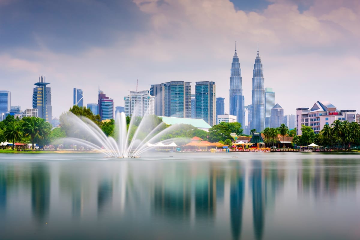 Skyline von Kuala Lumpur mit den Petronas Towers hinter einem Park, im Vordergrund ein Springbrunnen auf einem See