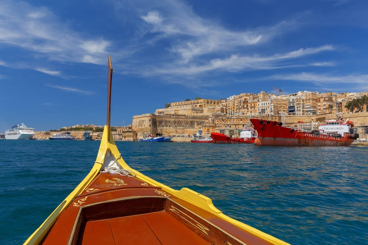 Blick über den bemalten Bug einer Dgħajsa auf die Bastionen und Sandsteinhäuser von Valletta, links ein Kreuzfahrtschiff am Kai
