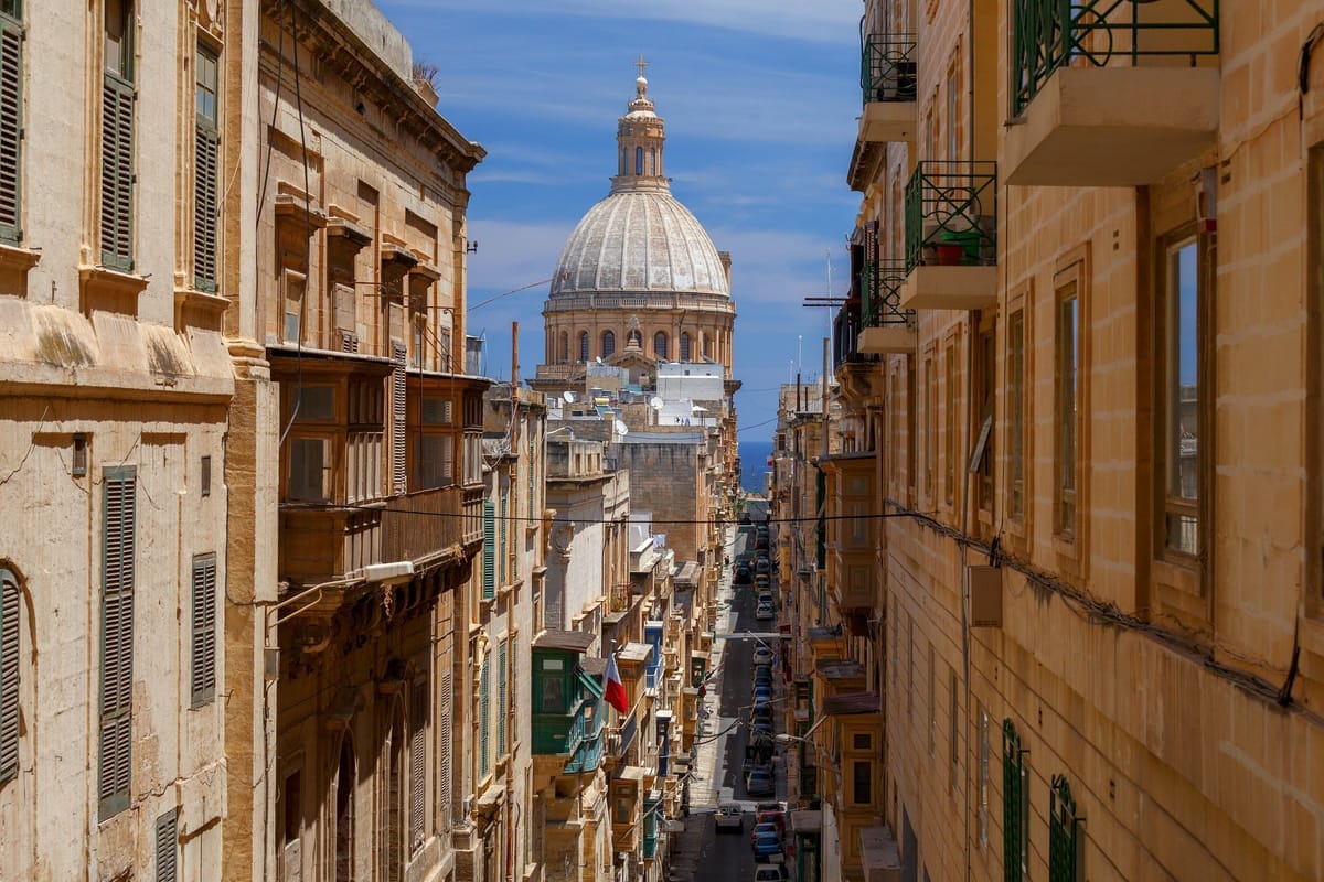 Enge Seitengasse in Valletta zwischen Sandsteinfassaden mit grünen Holzerkern, am Ende der Gasse die große Kuppel der Karmeliterkirche