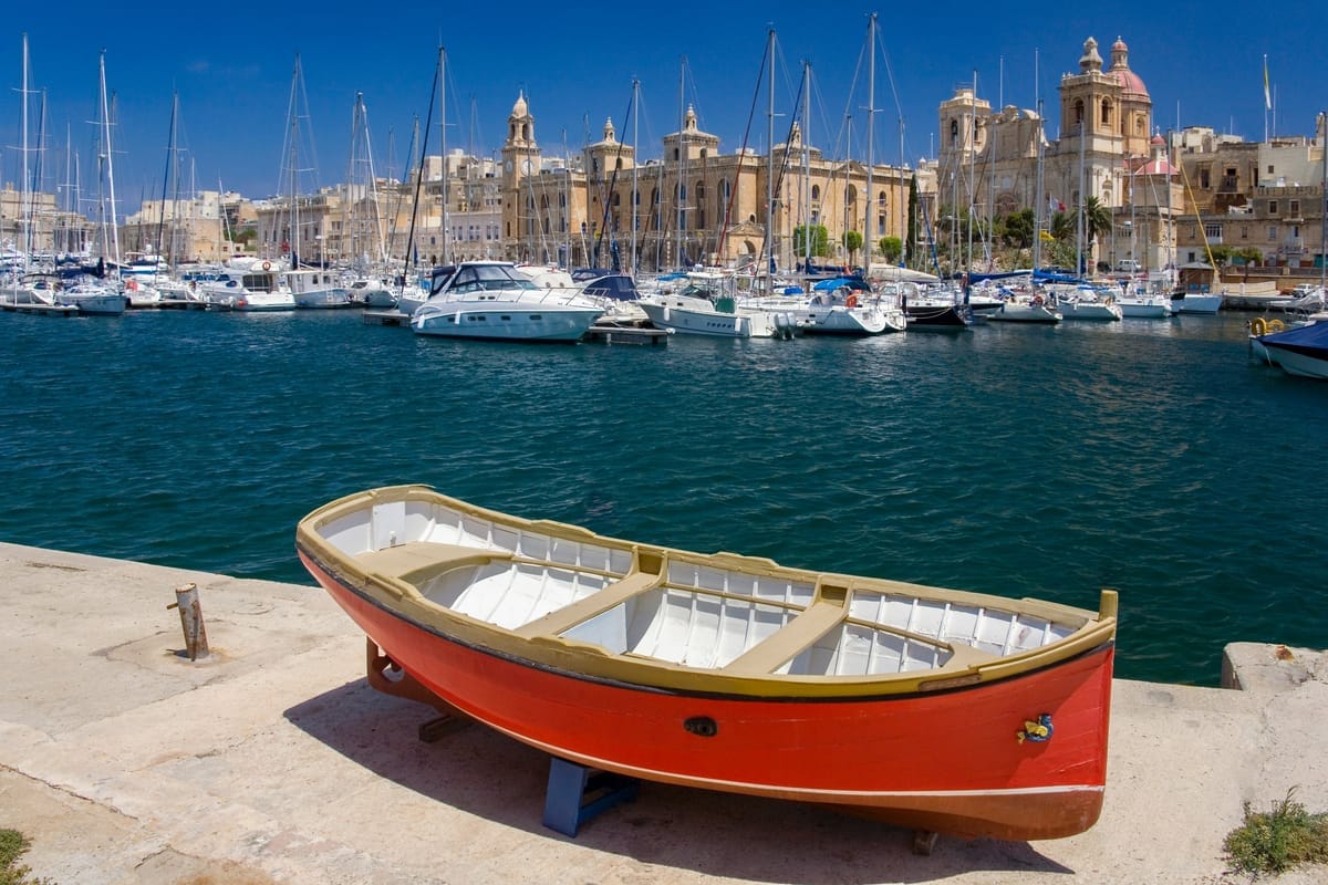 Ein rot-gelb bemaltes Holzboot liegt auf der Kaimauer der Marina von Vittoriosa, dahinter Yachten und die Häuserfront der Altstadt mit Kirchturm