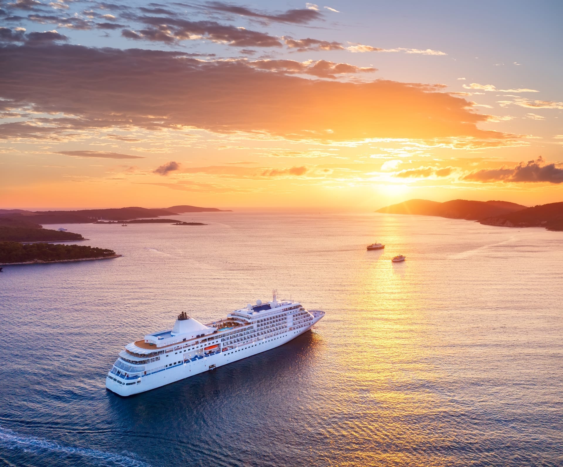 Kreuzfahrtschiff aus der Luft bei Sonnenuntergang vor einer Inselküste, die Sonne steht tief über dem Wasser
