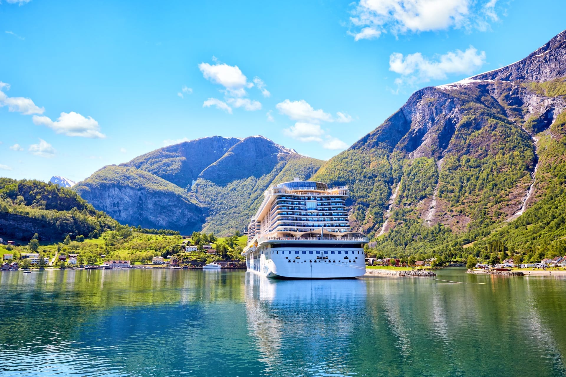 Kreuzfahrtschiff am Ende eines norwegischen Fjords, dahinter grüne Berghänge mit Schneeresten unter blauem Himmel