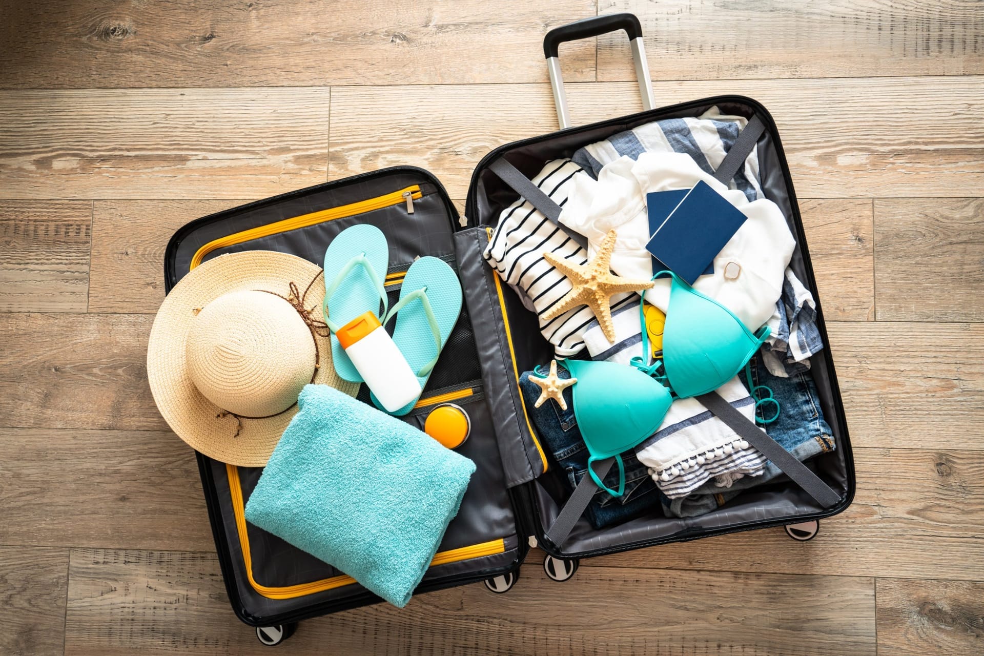 Geöffneter Koffer von oben, gepackt mit Sommerkleidung, Sonnenhut, Badeschuhen und Reisepass