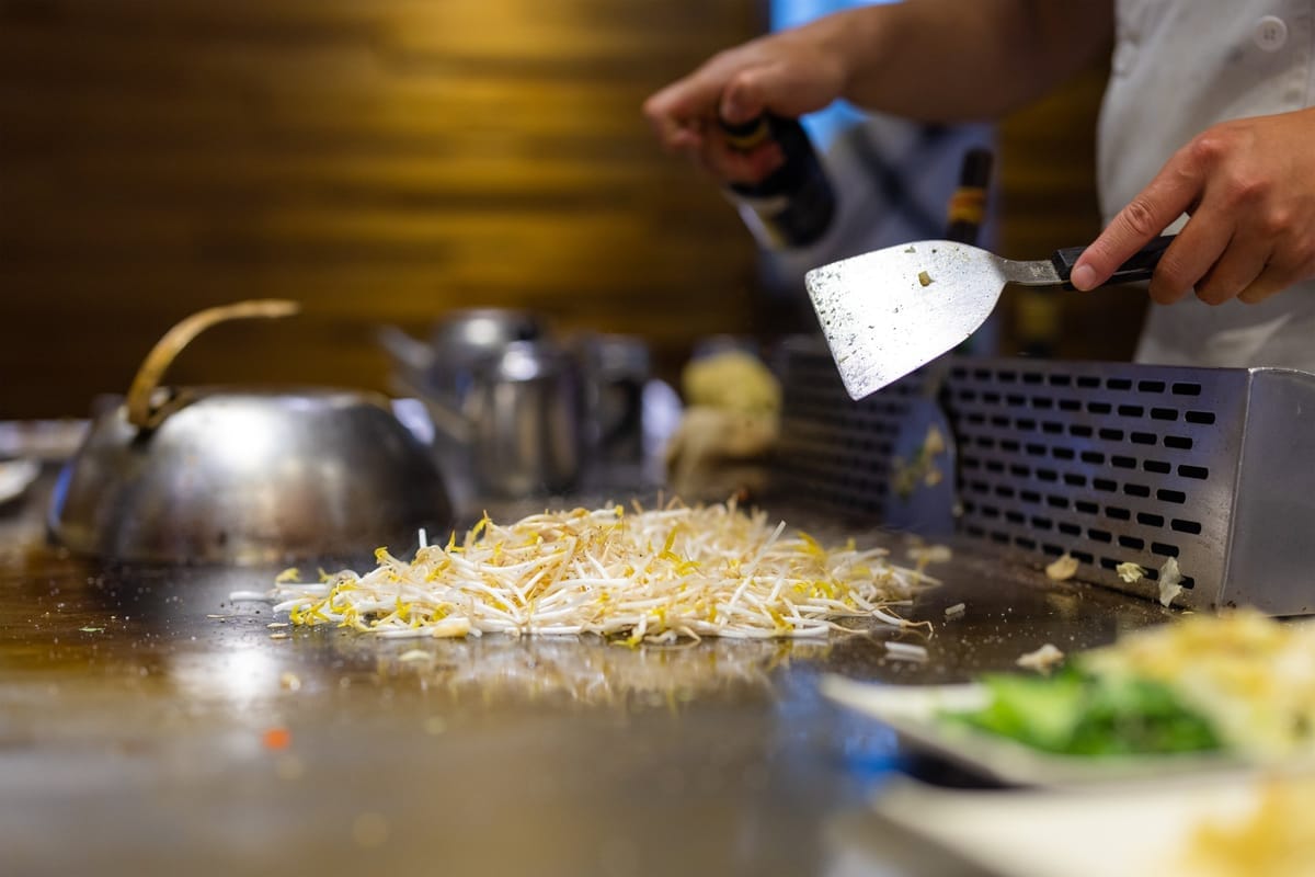 Koch brät Sojasprossen mit einem Pfannenwender auf einer heißen Teppanyaki-Grillplatte