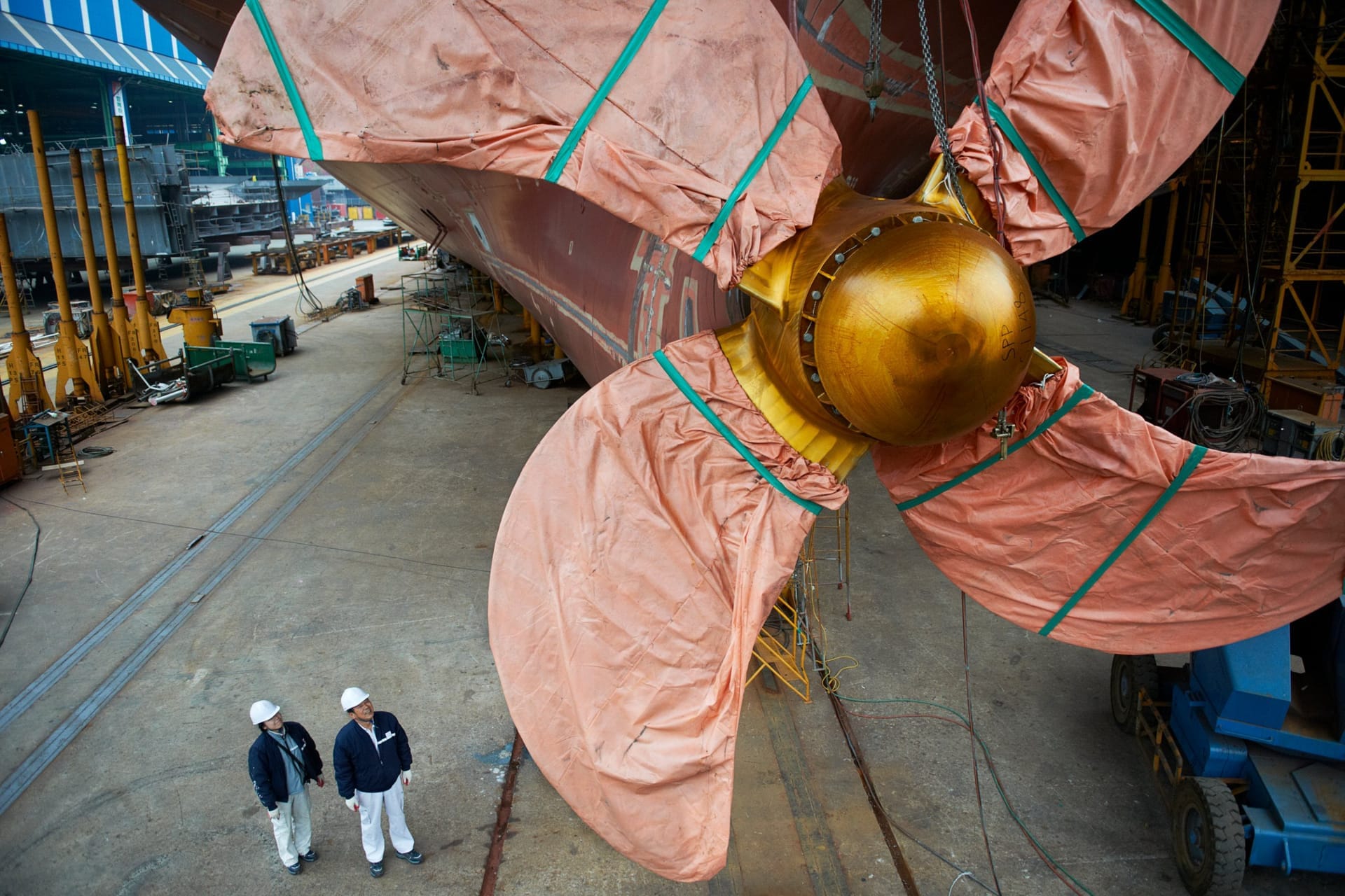 Riesiger Schiffspropeller mit vier verhüllten Flügeln unter dem Heck eines Schiffs im Trockendock, darunter zwei Werftarbeiter mit Helmen als Größenvergleich