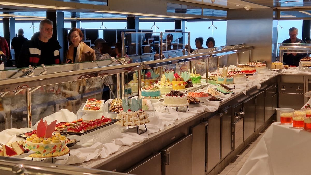 Lange Buffettheke mit zahlreichen Torten und Kuchen hinter einer Glasabdeckung, dahinter Panoramafenster mit Meerblick