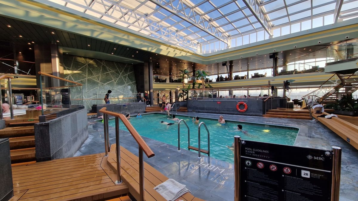 Pool unter einem großen Glasdach, im Vordergrund Holzdeck und ein Schild mit Poolregeln, daneben Whirlpools