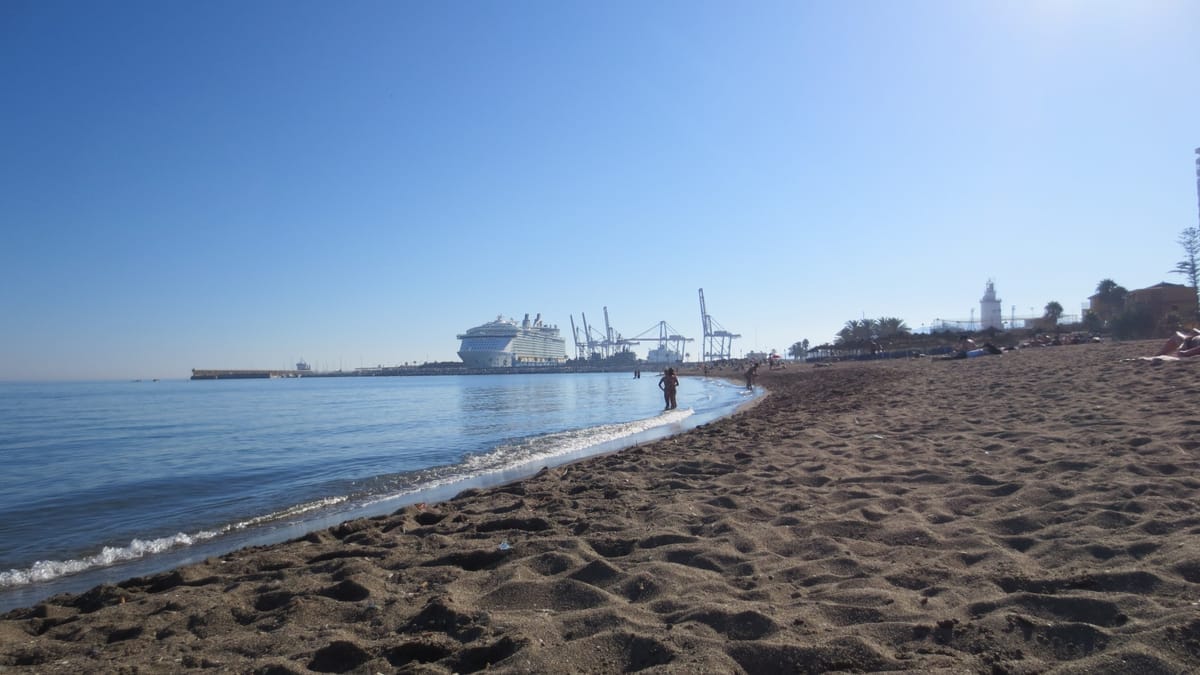 Sandstrand in Málaga mit ruhiger See, im Hintergrund Hafenkräne und die Oasis of the Seas