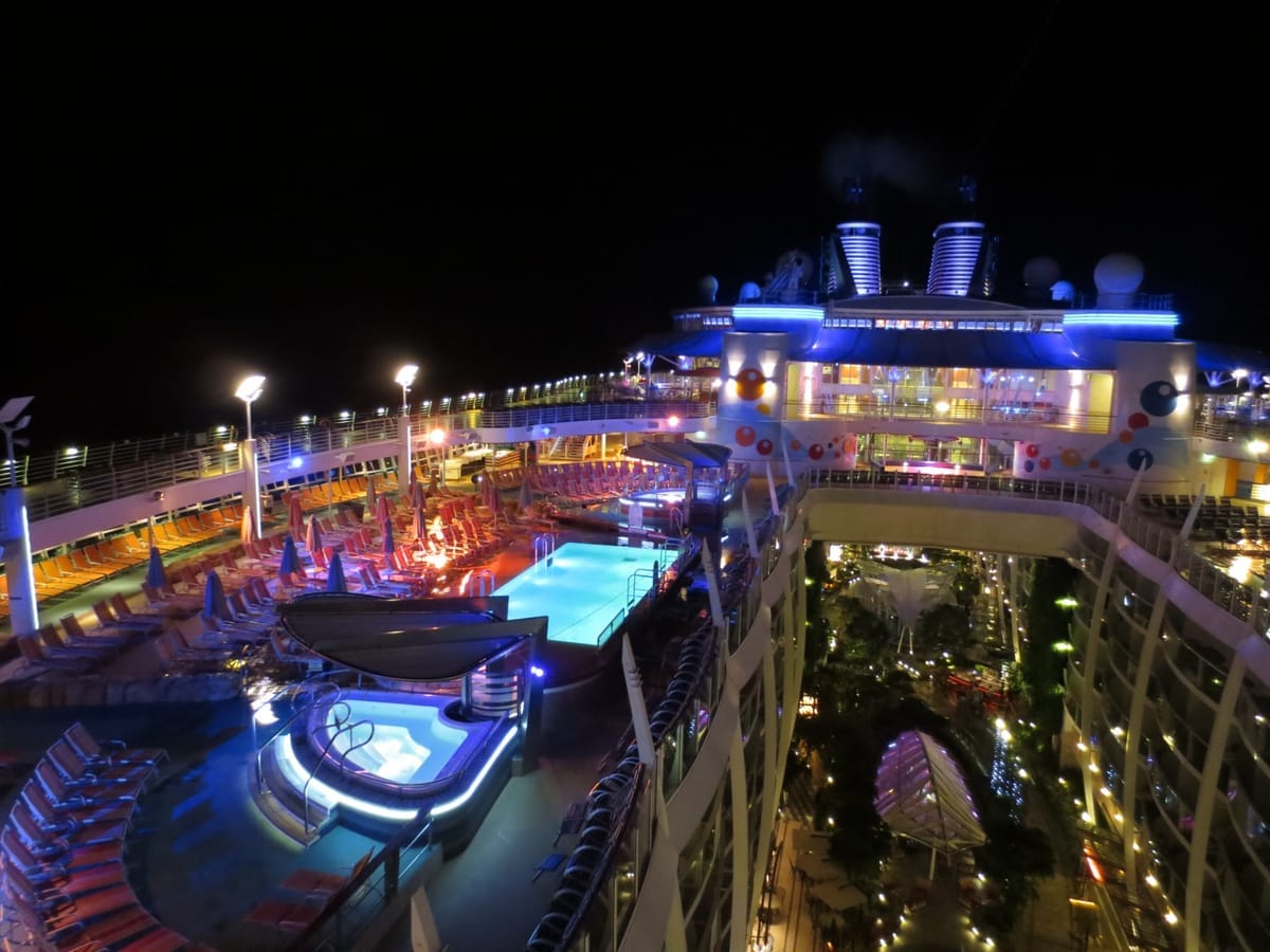 Beleuchtetes Pooldeck eines Kreuzfahrtschiffs bei Nacht, rechts der begrünte Innenhof zwischen den Kabinentrakten